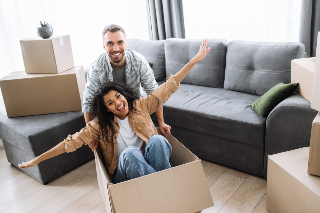 Happy married multiethnic couple at moving day. Cheerful husband with beautiful wife having fun with cardboard boxes while unpacking stuff. Satisfied woman and man smiling, excited about buying